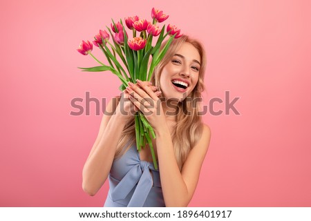 Happy Woman's Day. Excited young lady in cocktail dress holding bouquet of tulips on pink studio background. Cheerful blonde model with flowers as gift for spring holiday