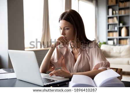 Close up serious focused woman looking at laptop screen, touching chin, sitting at desk, home office, thoughtful businesswoman pondering strategy, working on online project, searching information