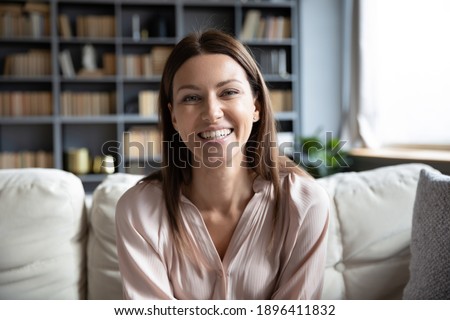 Head shot portrait smiling woman making video call to friends or relatives, using webcam, sitting on couch at home, happy young female blogger recording vlog, looking at camera, chatting online