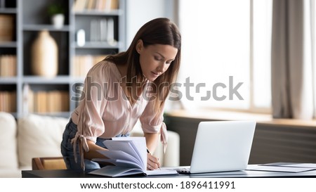 Confident businesswoman writing in notebook, standing at work desk, home office, young female planning workday, meetings in organizer, student making notes in copybook, watching webinar, lecture Royalty-Free Stock Photo #1896411991