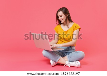 Full length portrait of beautiful smiling attractive young brunette woman 20s wearing yellow casual t-shirt sitting on floor working on laptop pc computer isolated on pink color background studio