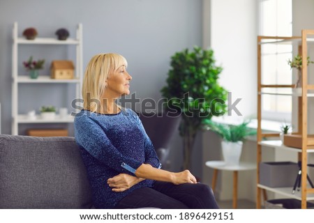 Quiet moment. Calm pensive smiling senior woman looking out the window sitting on sofa in cozy living room. Pensioner enjoys a lazy weekend or vacation. Woman sits waiting for her grandchildren.
