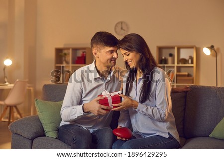 Young man tenderly looks at his girlfriend giving her a gift for Valentine's Day. Loving couple exchanging gifts sitting on the couch at home in a cozy room. Valentine's Day concept.