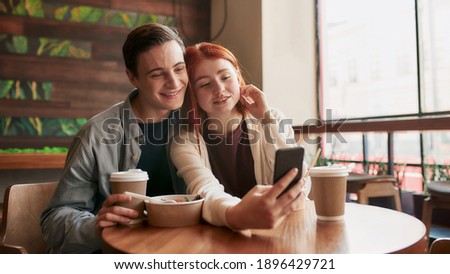 Happy teenage couple smiling while taking selfies using smartphone, sitting in a cafe together on a daytime. Modern relationships concept. Selective focus. Web Banner