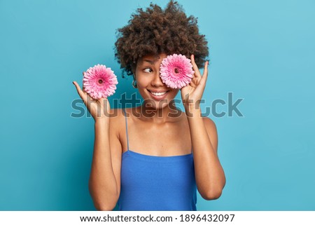 Positive dark skinned young woman covers eyes with pink gerberas smiles happily has curly bushy hair dressed in casual clothes arranges bouquet isolated over blue background. Floral concept.
