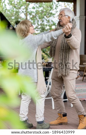 Affectionate senior couple in warm stylish casualwear dancing on wooden floor of patio by their country house against served table