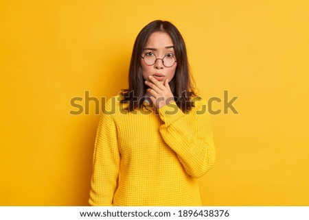 Portrait of worried surprised Asian woman holds chin looks concerned at camera wears transparent glasses and sweater isolated over yellow background. People face expressions and reaction concept Royalty-Free Stock Photo #1896438376