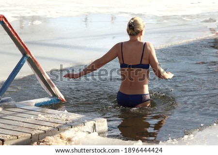 Winter swimming sport, a young European woman in a swimsuit enters the ice hole water on a Sunny frosty winter day, healthy lifestyle