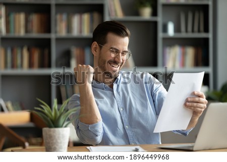 Emotional young businessman in eyeglasses reading paper notification, feeling excited of getting unbelievable news. Happy overjoyed man getting dream job offer, celebrating success in office. Royalty-Free Stock Photo #1896449968