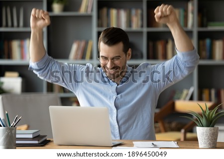 Overjoyed young man in glasses looking at laptop screen, celebrating online lottery win or reading unbelievable news at office. Emotional millennial guy getting dream job offer, feeling joyful at home Royalty-Free Stock Photo #1896450049