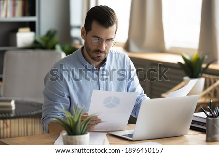 Concentrated young businessman ceo manager in eyewear looking at marketing research report, analyzing statistics data in charts, developing growth strategy, working on computer alone in modern office. Royalty-Free Stock Photo #1896450157
