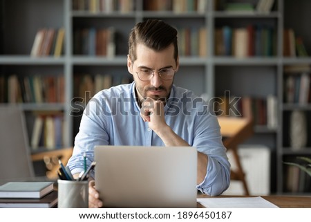 Thoughtful young 30s caucasian businessman in eyeglasses looking at computer screen, working distantly online at home office, solving problems, making difficult decision, freelance workday concept. Royalty-Free Stock Photo #1896450187