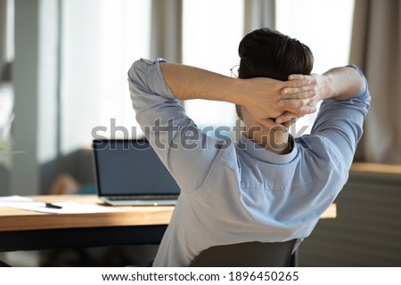 Back rear view happy young male worker employee resting on chair with folded hands behind head, enjoying break pause time during difficult workday in modern office, feeling relaxed alone indoors. Royalty-Free Stock Photo #1896450265
