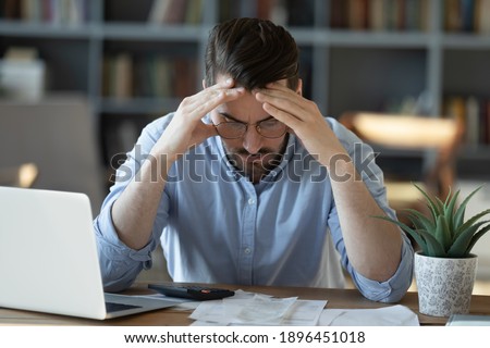 Frustrated young man in glasses sitting at table, feeling stressed calculating earnings or domestic expenses. Unhappy millennial guy suffering from lack of money, banking debt bankruptcy concept. Royalty-Free Stock Photo #1896451018