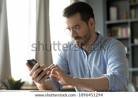 Happy smiling millennial man in eyeglasses looking at smartphone screen, enjoying using mobile software applications, web surfing information or communicating with client online at home office.
