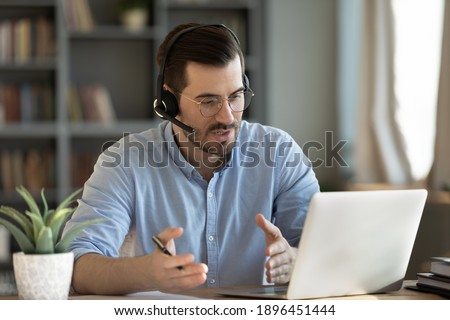 Concentrated skilled millennial caucasian businessman in glasses wearing headphones with mic, taking part in online web camera negotiations meeting using computer app, distant communication concept. Royalty-Free Stock Photo #1896451444
