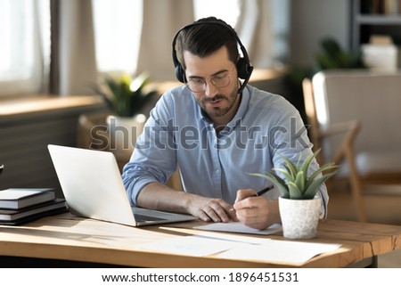 Focused young businessman in eyeglasses wearing wireless headset with microphone, involved in online video call negotiations meeting with partners colleagues or studying distantly, writing notes. Royalty-Free Stock Photo #1896451531