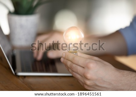 Close up cropped young man holding lighting bulb in hands, working on startup project on computer, having brilliant creative idea, feeling motivated and inspired at workplace, business success concept Royalty-Free Stock Photo #1896451561
