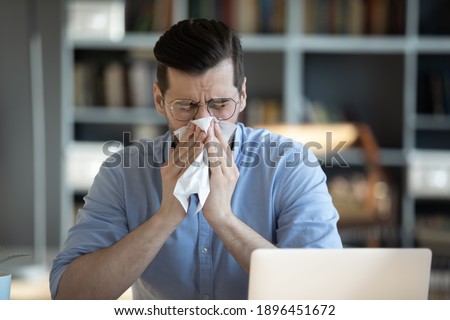 Sick millennial unhappy businessman wiping runny nose with handkerchief, suffering from seasonal allergic reaction or sneezing, caught cold feeling first flu symptoms sitting alone at workplace. Royalty-Free Stock Photo #1896451672