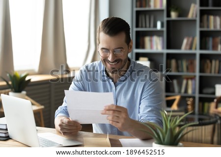 Smiling young businessman in eyeglasses looking at paper correspondence, reading pleasant good news. Excited happy male entrepreneur getting bank loan approvement notification at home office. Royalty-Free Stock Photo #1896452515