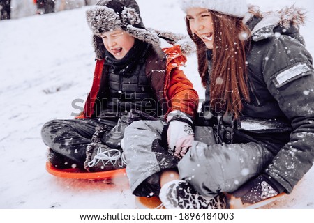 one boy and one girl with long hair on the street in the afternoon in winter a lot of snow rolling downhill on a round red board sitting laughing in warm clothes snow falls on the face