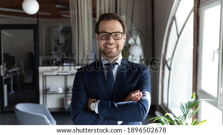 Head shot smiling confident young businessman executive wearing glasses and suit standing in modern office room, happy successful entrepreneur employee intern with arms crossed looking at camera