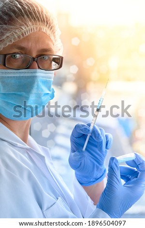 Medic, doctor or nurse with coronavirus vaccine vial and syringe. Mature doctor in face mask, glasses, hat, blue gloves and protective white gown. Romantic view over town with sunset behind