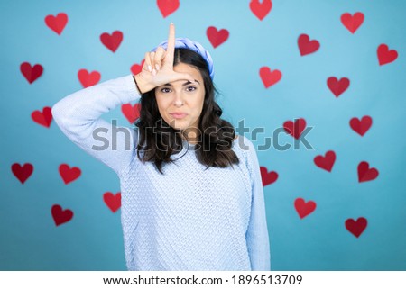 Young beautiful woman over blue background with red hearts making fun of people with fingers on forehead doing loser gesture mocking and insulting.