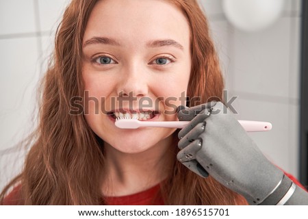 Portrait view of the girl with prosthesis hand looking to the camera and brushes teeth with a dental brush to prevent tooth decay. Dental and healthy tooth concept. Stock photo