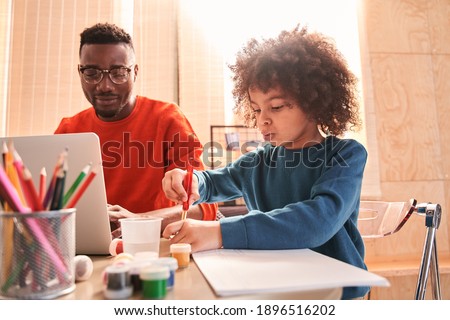 Cute pensive curly boy pondering what to drawing while sitting over a blank sheet of paper at the wooden table. Boy spending time near his father working with laptop at kitchen. Stock photo