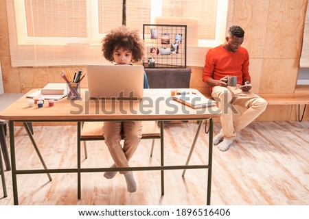 Smart curly boy is thinking about new hometask while sitting at online lesson in front of a laptop at the kitchen. His multiracial father working at the background. Stock photo