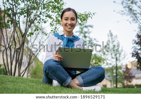 Portrait of young beautiful asian student studying online, sitting on grass. Distance learning concept. Smiling woman using laptop computer, working freelance project outdoors