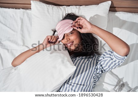 Cute brunette multiracial woman laying in her bed in striped pajamas and sleep mask, top view, Girl looking flirty at the camera and relaxing. Stock photo