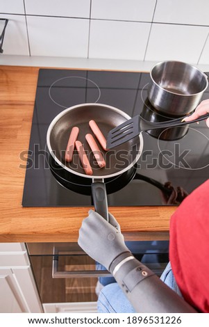 Lovely serene woman with prosthesis hand frying sausages at the pan while cooking food in kitchen at home. Stock photo. Disabled people concept
