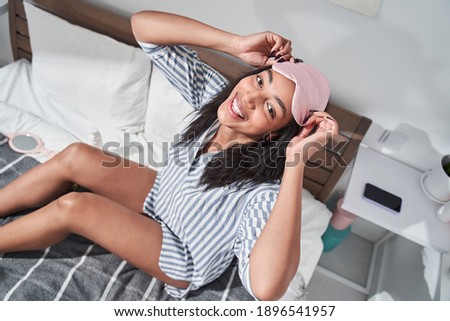 Top view of the attractive multiracial woman adjusting sleep mask at her head while sitting at the bedroom and taking care of her skin. Stock photo