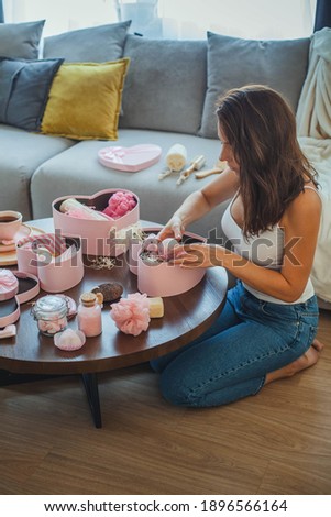 Young beautiful woman opens pink colored heart shaped gift boxes with homemade bath supplies, and body care cosmetics products for woman on white marble background. Concept of Valentines day gifts.