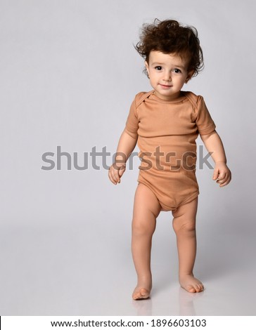 Child learns to walk. Cute smiling little curly baby boy stands on a gray background in the studio and looks at the camera. Concept of new children's skills. Place for text.