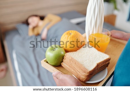 Close-up image of man bringing tray with sandwich, orange juice and fruits in bed where his wife is sleeping on Valentines day morning