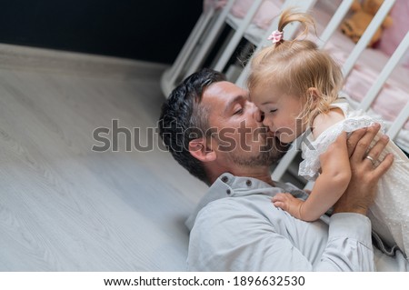 The Caucasian man lies on his back and holds his daughter above him in the children's room