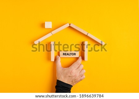 Male hand placing realtor sign in a house made of wooden blocks on yellow background.