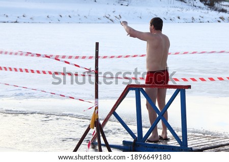 Winter swimming sport, a Caucasian man in a swimming trunks stand up near the ice hole water on planked footway on a Sunny frosty winter day, healthy lifestyle