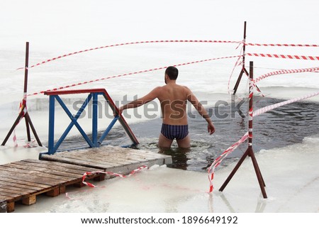 Winter swimming sport, a Caucasian man in a swimming trunks enters in the ice hole water on planked footway on a Sunny frosty winter day, healthy lifestyle