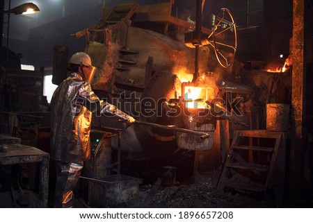Hardworking in foundry. Workman in temperature protection suit pouring liquid metal into the bucket. Steel production. Royalty-Free Stock Photo #1896657208