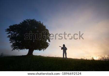 Horizontal snapshot of two silhouettes on purple sky background, of a spaceman in space suit playing guitar and a big old tree on the right. Concept of music, cosmonautics and nature.