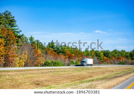 Powerful gray big rig industrial diesel semi truck with refrigerator semi trailer transporting cargo running on the divided highway road with colorful maples autumn trees in New England