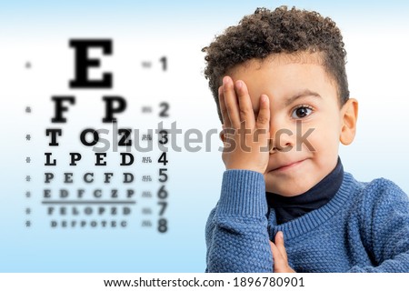 Close up face shot of little Afro American boy testing vision. Kid with closing on eye with hand. Vision chart with block letters and focus point in background.