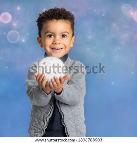 Close up conceptual portrait of cute afro american boy holding the moon in hands. Kid with happy surprised facial expression against cosmos background. 