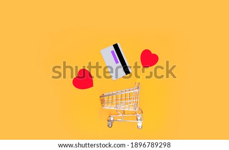 Shopping basket and white credit card with red hearts in flight on a bright yellow background. Valentine's day shopping concept. copy space. view from above.