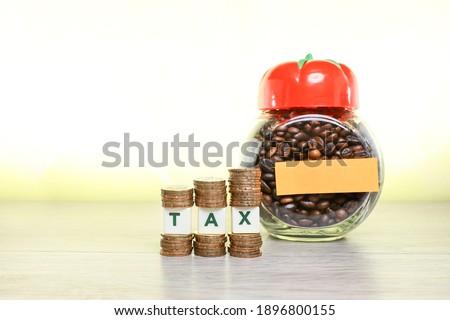 Block letters on tax sandwiched between staked of coins with a glass jar full with coffee beans 