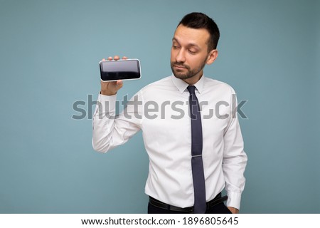 Photo of handsome good looking brunet man with beard wearing casual white shirt and tie isolated on blue background with empty space holding in hand and showing mobile phone with empty screen for